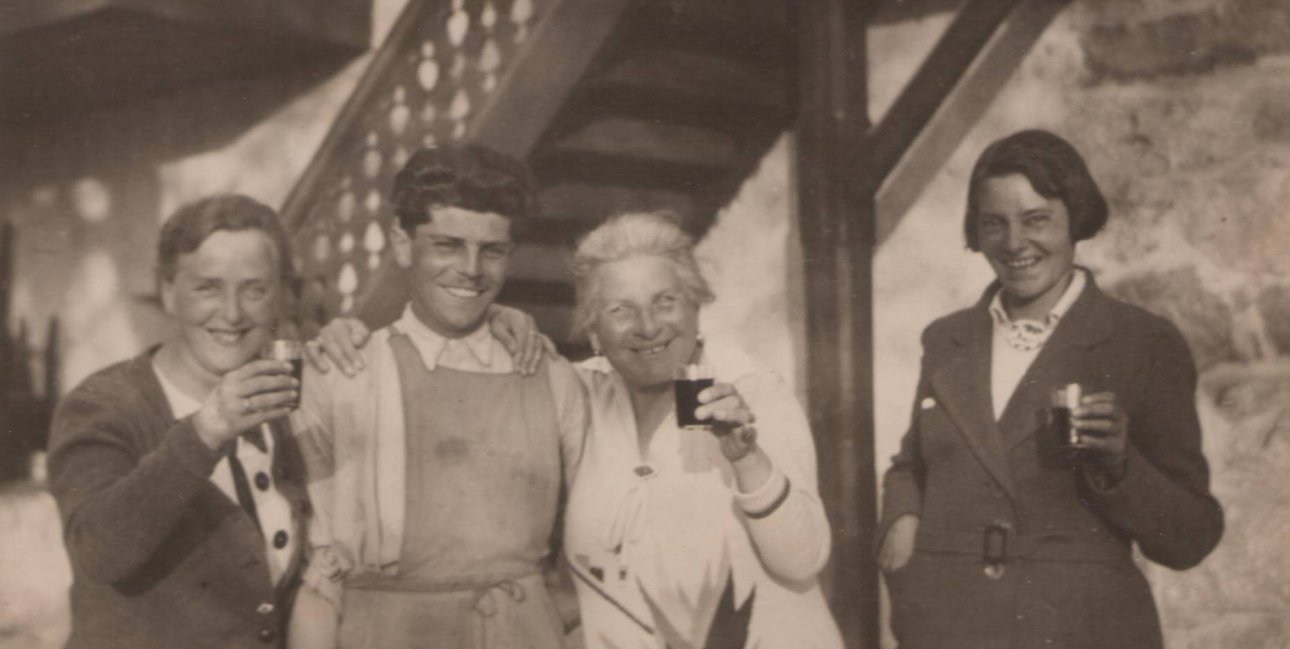 Ein historisches Schwarz-Weiß-Foto von vier Personen, die lächelnd in die Kamera schauen und mit Gläsern in der Hand anstoßen. Im Hintergrund ist eine rustikale Holztreppe zu sehen.