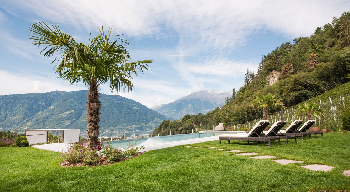 Infinity Pool mit Palme, Liegestühlen und Blick auf Meran & Umgebung im Wellnesshotel Kiendl in Schenna