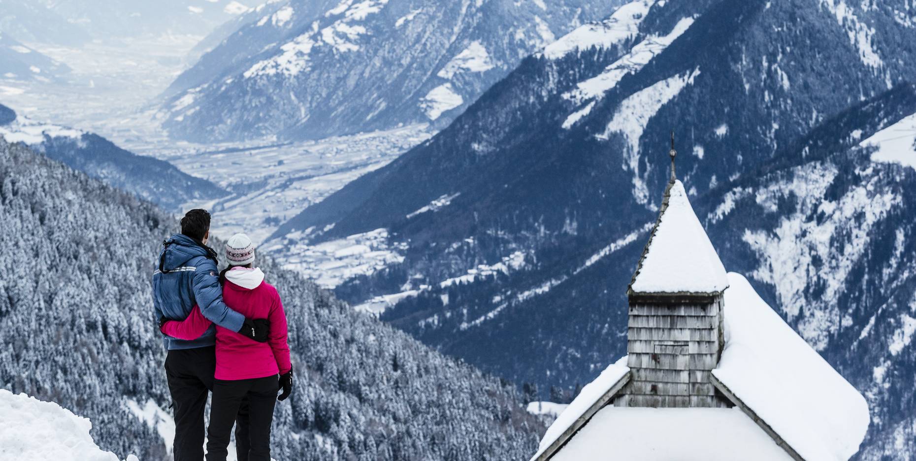 Ein Paar in Winterkleidung umarmt sich vor einem großen Bild verschneiter Berge.