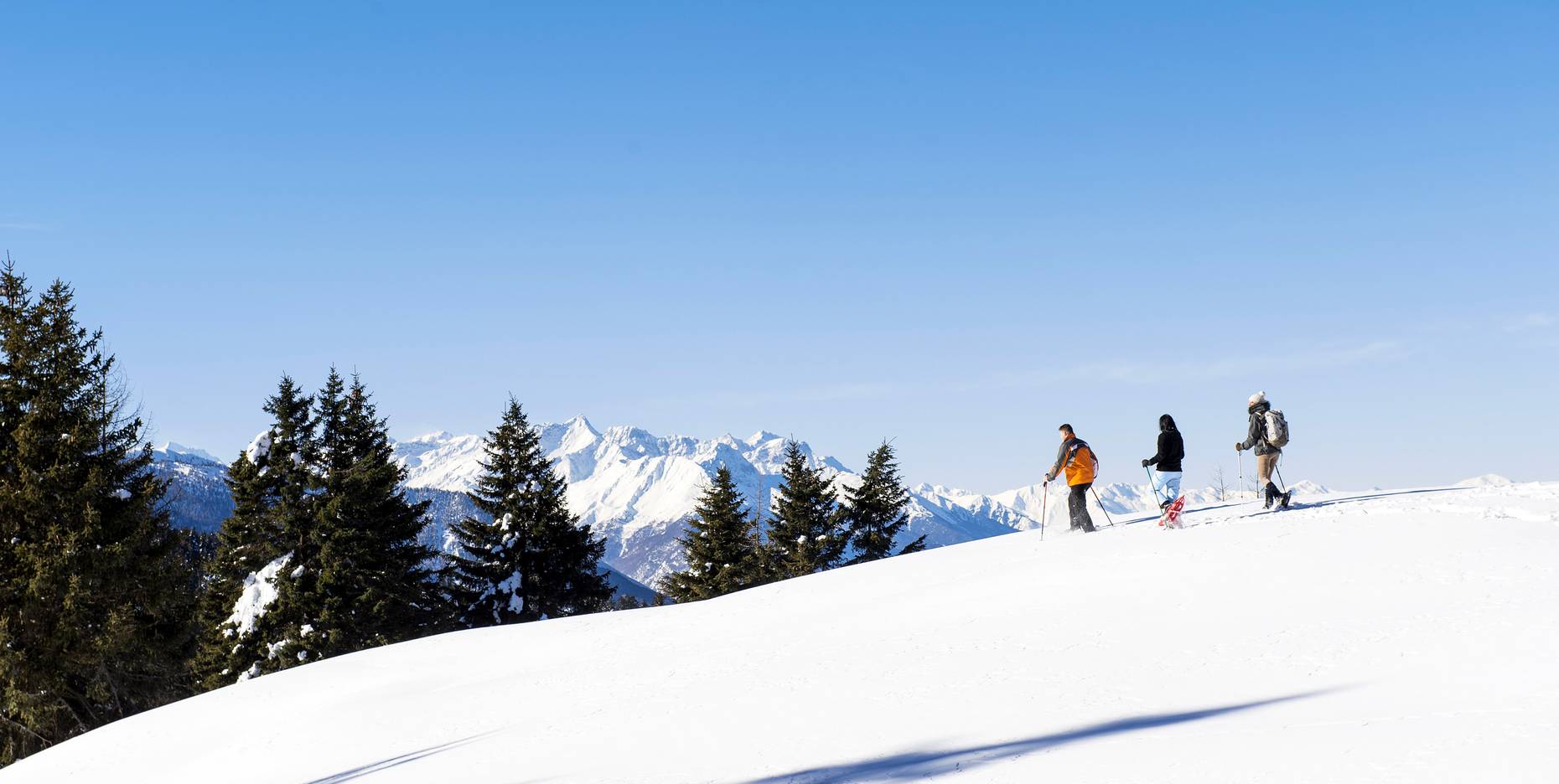 Drei Personen beim Schneeschuhwandern auf einer verschneiten Bergkette unter strahlend blauem Himmel.