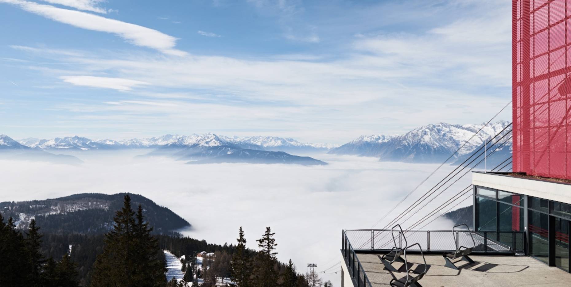 Eine Aussichtsterrasse mit Liegestühlen und einem auffälligen roten Gebäude vor einem Panorama aus verschneiten Bergen und einer dichten Wolkendecke im Tal.