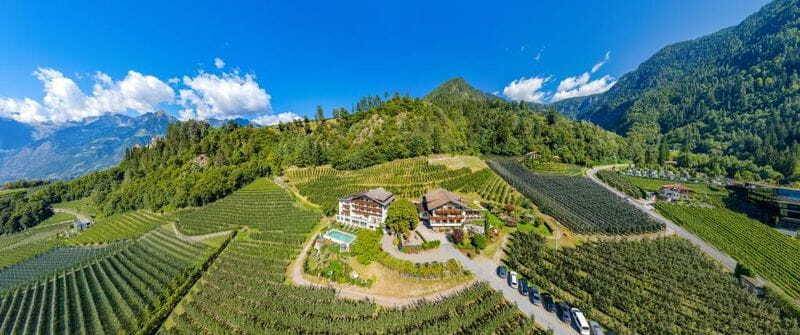 Luftaufnahme eines Hotels, umgeben von weitläufigen Obstplantagen in einer bergigen Landschaft bei strahlend blauem Himmel.