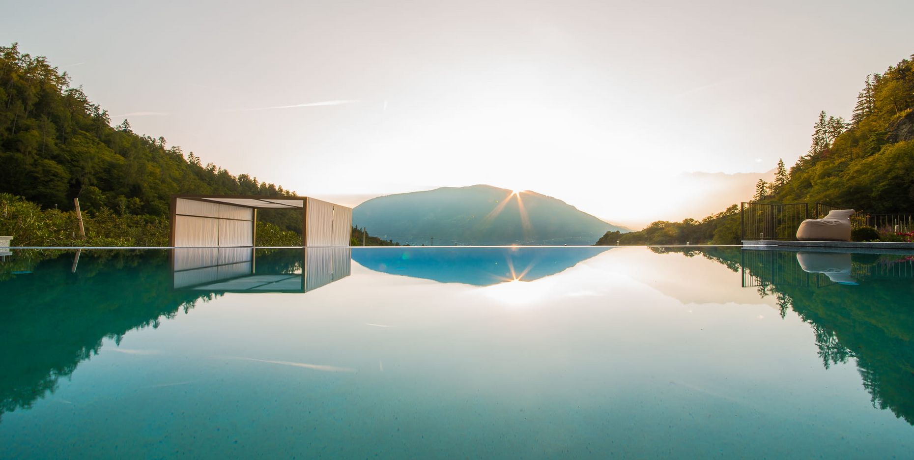 Ein Moment der vollkommenen Stille: Der magische Sonnenuntergang spiegelt sich im spiegelglatten Wasser des Infinity-Pools. Ein Infinity-Pool spritzt die umliegenden Berge und den Sonnenuntergang wider, mit einer schlichten Pool-Lounge am linken Rand.
