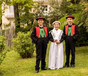 Drei Kinder stehen in einem Garten: Zwei Jungen tragen traditionelle schwarze Trachten mit roten Westen und Hüten, ein Mädchen trägt ein weißes Kleid mit Blumenkranz.