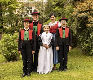 Eine fünfköpfige Familie posiert in traditioneller Tracht auf einer Wiese vor einem begrünten Haus.