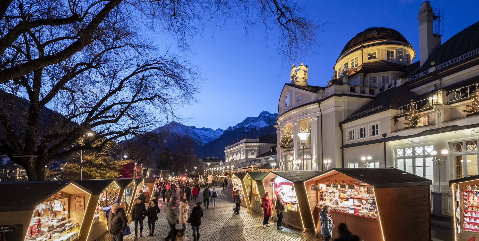 Nächtliche Einkaufsstraße mit beleuchteten Geschäften, Passanten, kahlen Bäumen und einer Bergsilhouette im Hintergrund.