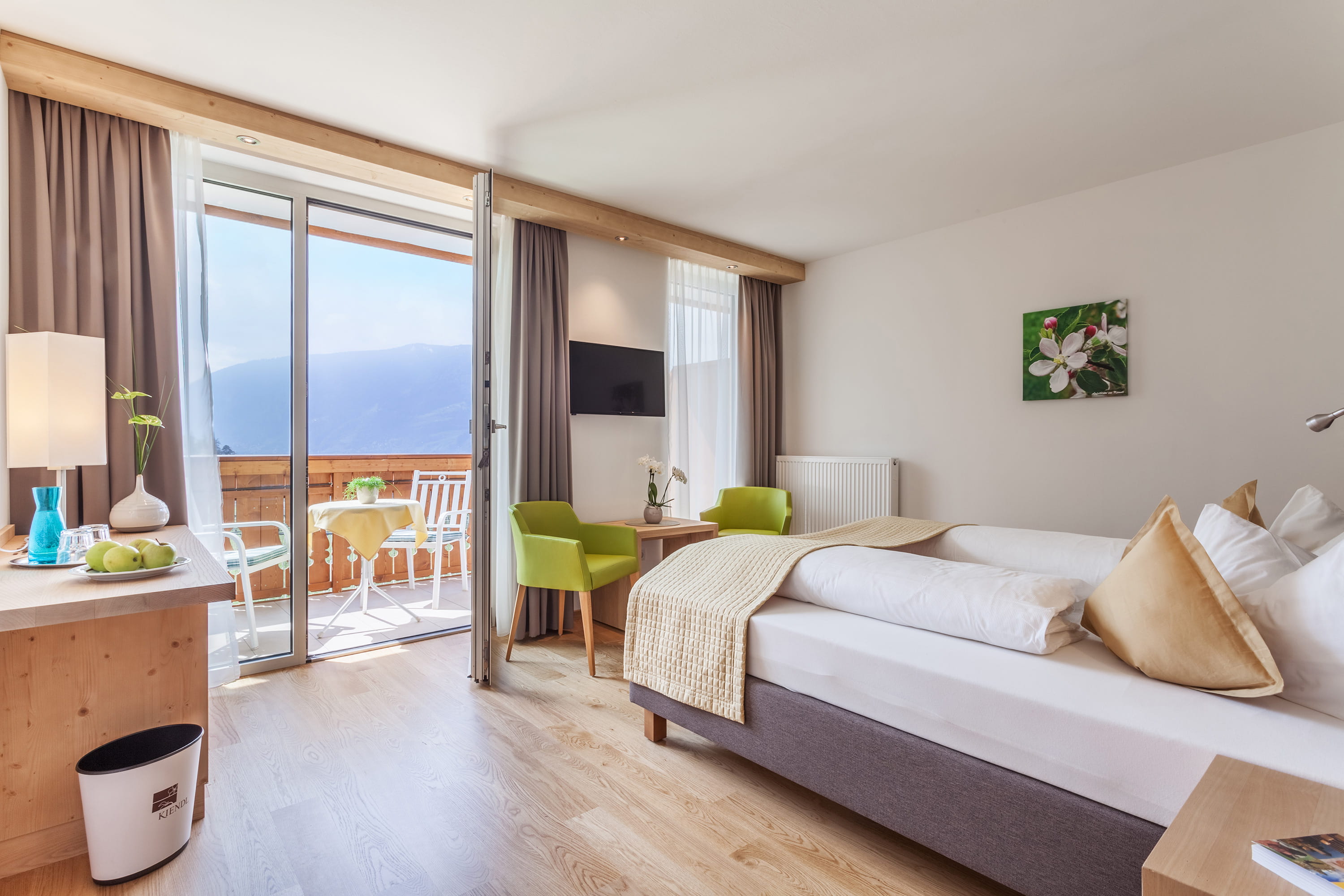 Ein helles Hotelzimmer mit einem Doppelbett, zwei grünen Sesseln und einer Terrassentür, die den Blick auf die Berge freigibt.