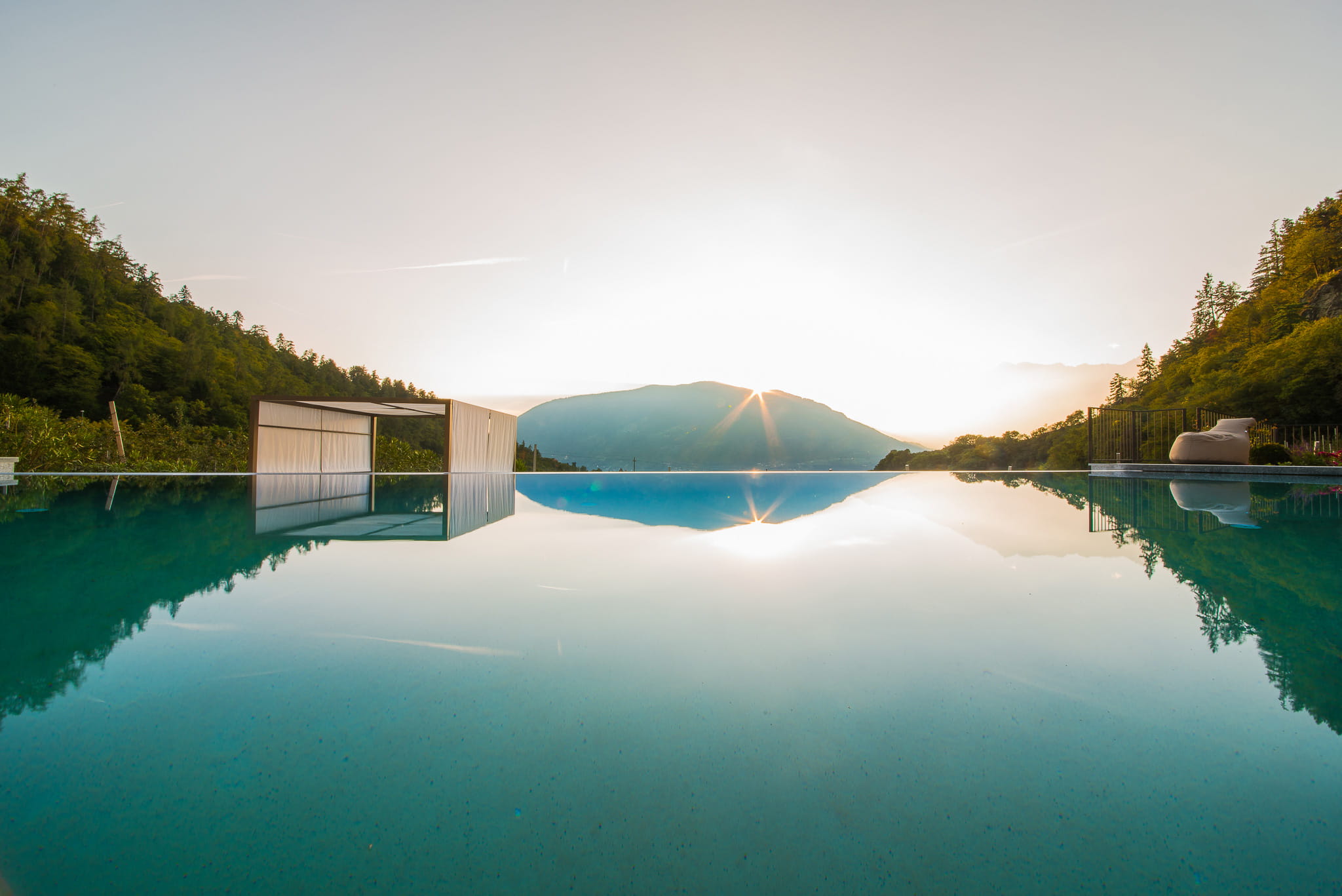 Ein Infinity-Pool spiegelt bei Sonnenuntergang das umliegende Bergpanorama und eine weiße Pool-Cabana wider.